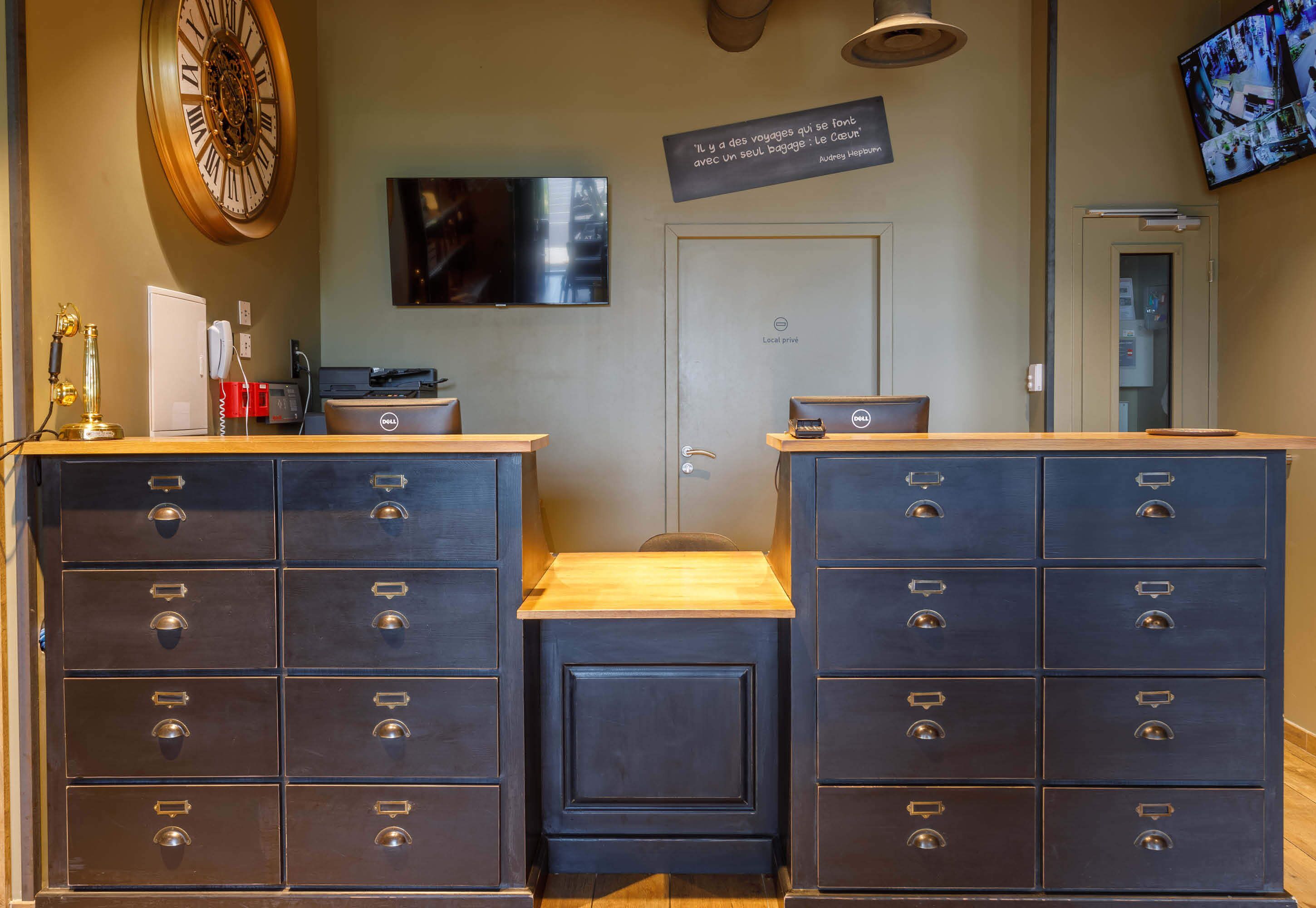 A hotel reception area with a large wooden clock, a flat-screen TV, and a sign with a quote. There are two dark blue filing cabinets and a small wooden table in the center.