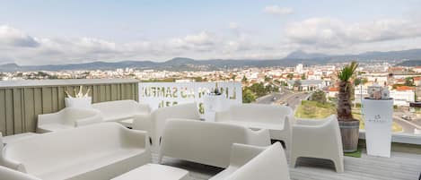 A rooftop terrace with white plastic furniture, a view of a cityscape, and a mountain range in the distance.