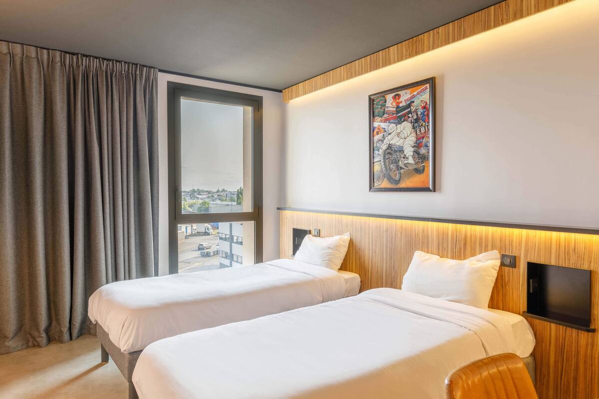 A hotel room with two beds, a large window with curtains, a mounted TV, and a wall-mounted artwork.