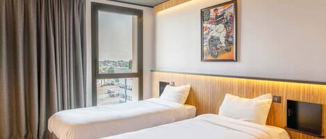 A hotel room with two beds, a large window with curtains, a mounted TV, and a wall-mounted artwork.