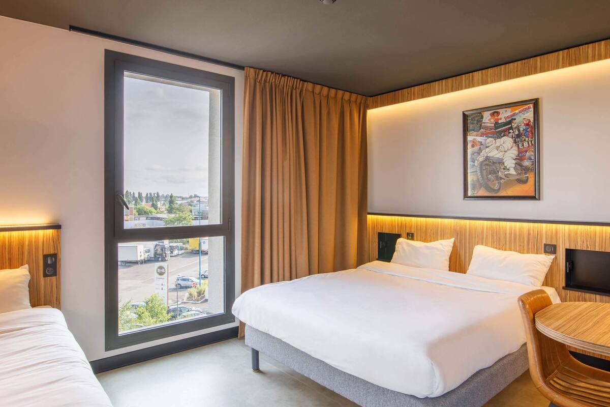 A hotel room with a large bed, a desk, a chair, a window with curtains, and a wall-mounted TV.