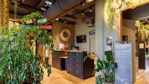A reception area with a wooden desk, a large clock, and a chalkboard with writing. There are plants and a sign with city names.