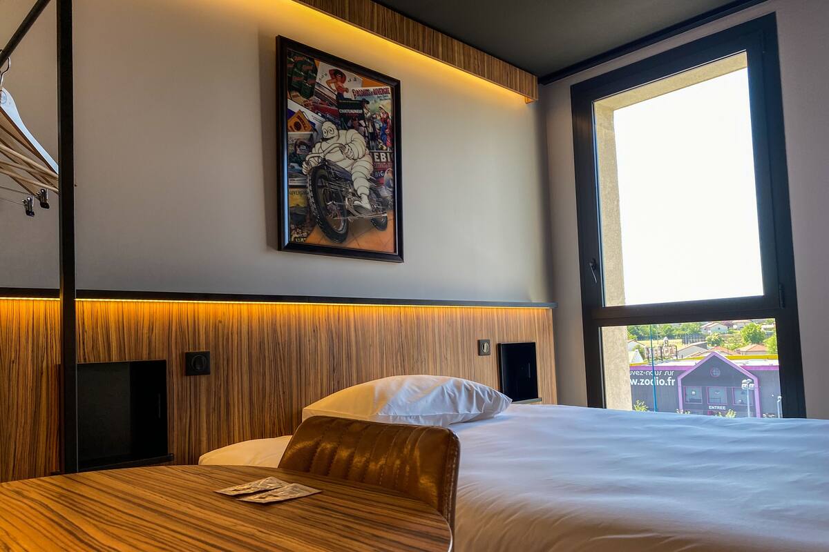 A hotel room with a large window, a bed with white bedding, a wooden headboard, and a framed poster on the wall.