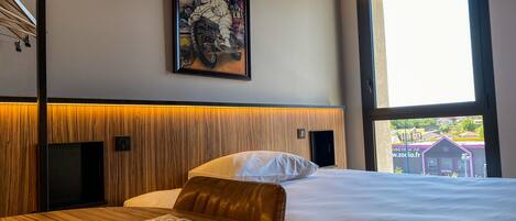 A hotel room with a large window, a bed with white bedding, a wooden headboard, and a framed poster on the wall.