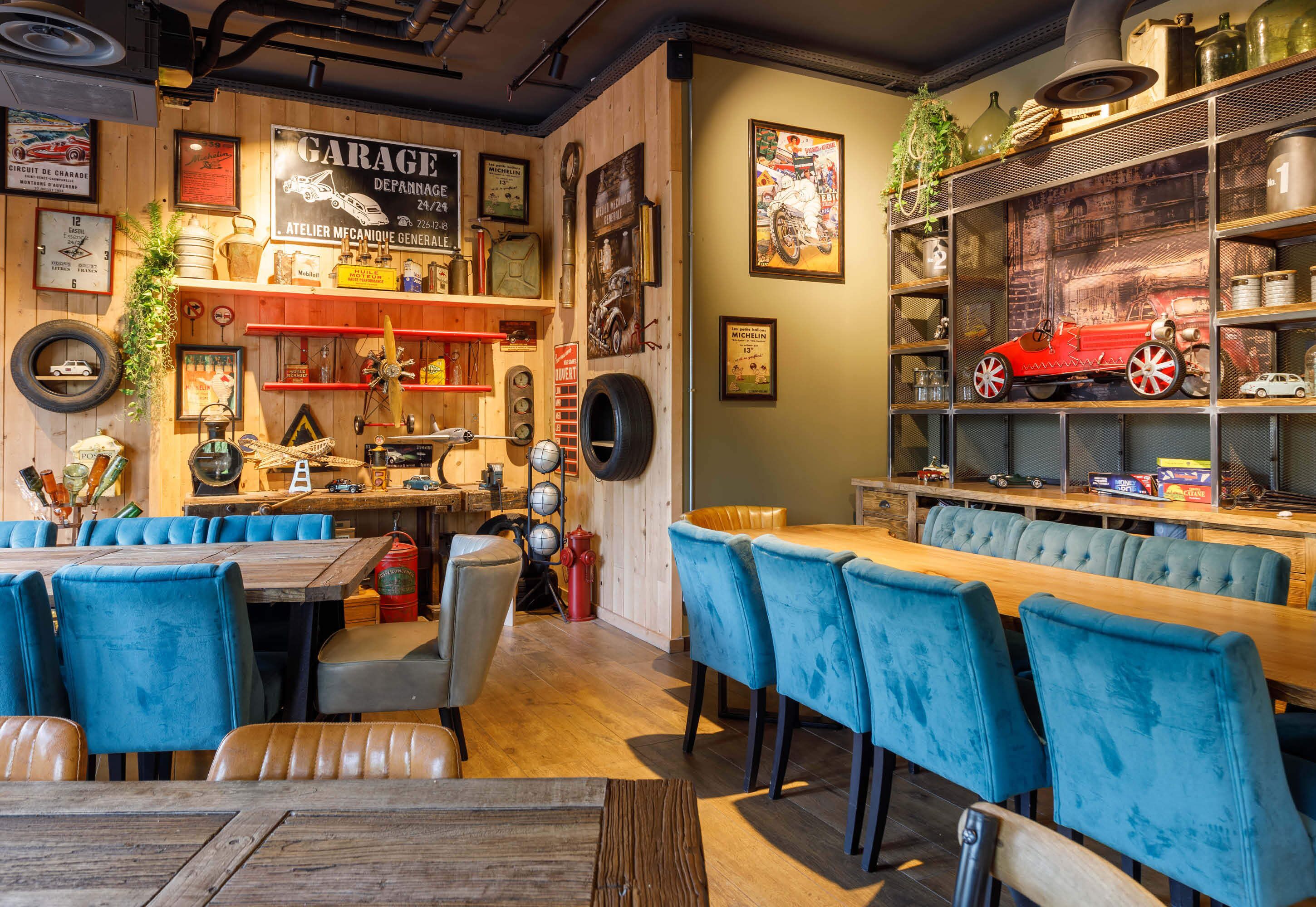 Un restaurante con mesas de madera y sillas azules, una pared con temática de garaje, y estantes con coches a escala y objetos antiguos.