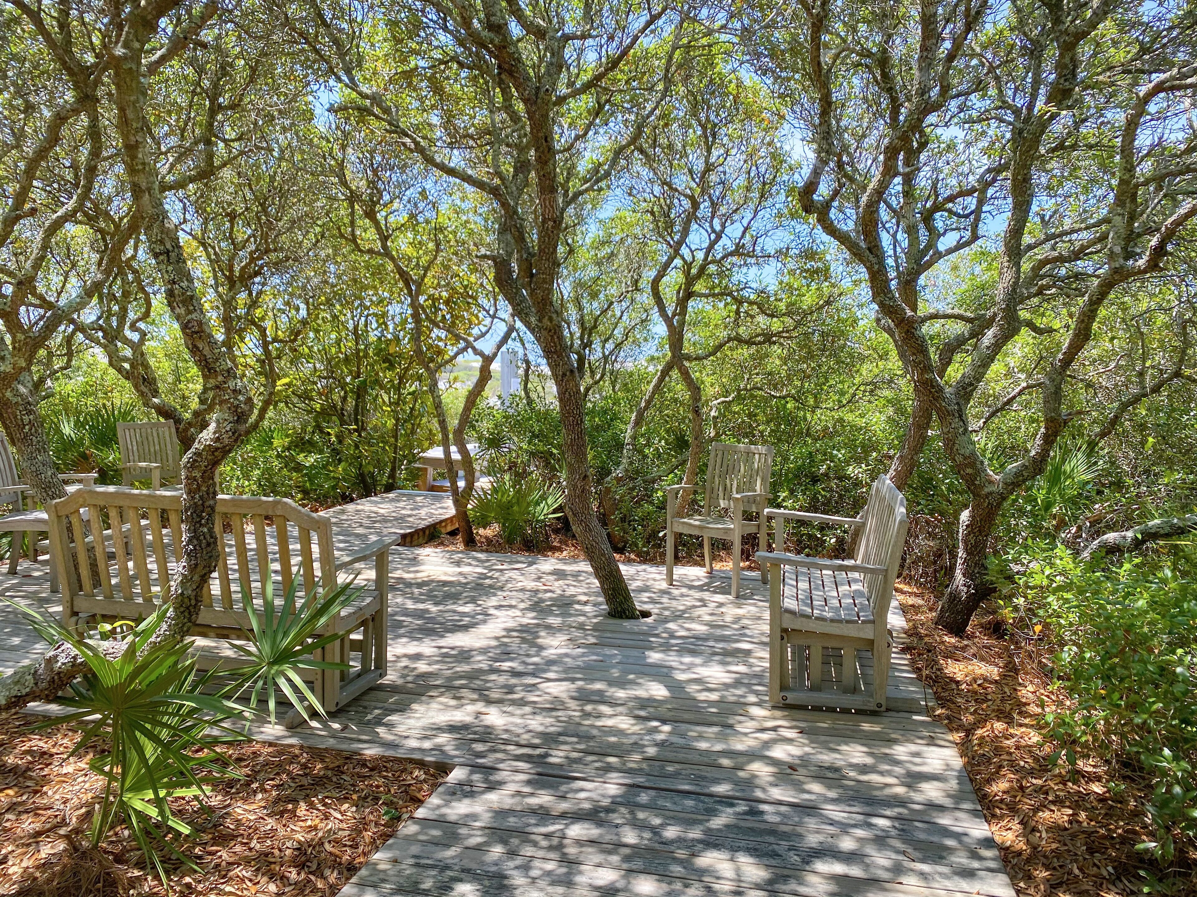 A wooden deck with chairs and benches, surrounded by trees and greenery.