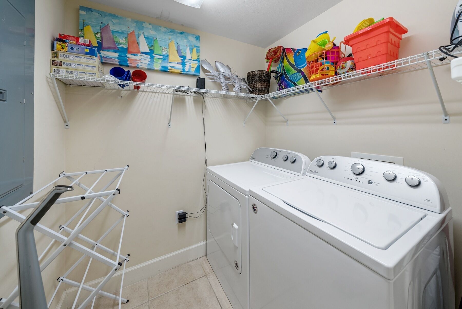 Un cuarto de lavandería con lavadora y secadora blancas, repisas con diversos objetos y un tendedero.