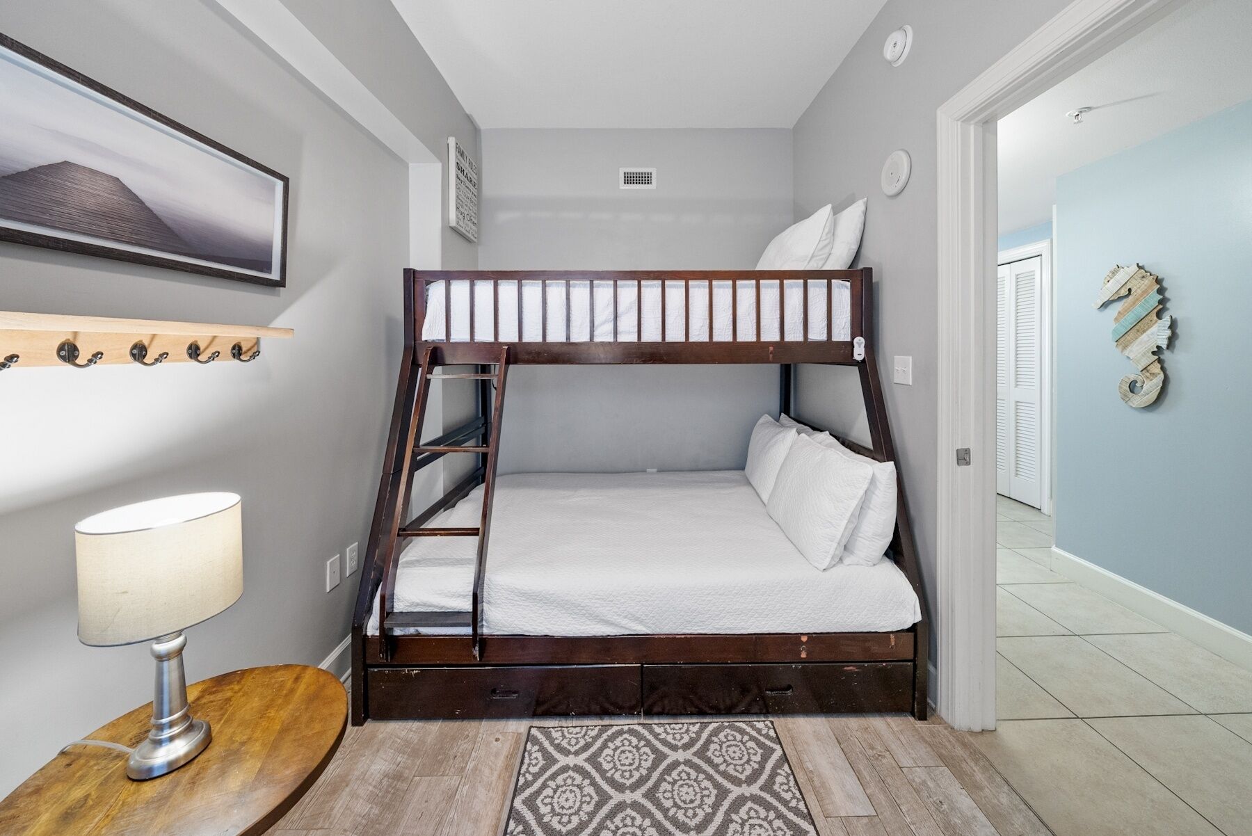 A bunk bed with a wooden frame and white bedding, a bedside table with a lamp, a patterned rug, and a wall-mounted shelf with hooks.