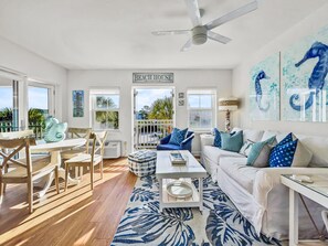 A spacious living area with a dining table, chairs, a sofa, and a coffee table. A sign reading "BEACH HOUSE" is visible, indicating a beach-themed decor. - Golden On 30a 1 Bedroom Condo by RedAwning (Santa Rosa Beach)