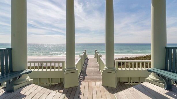 A covered wooden deck with white columns and benches, overlooking a beach and ocean.