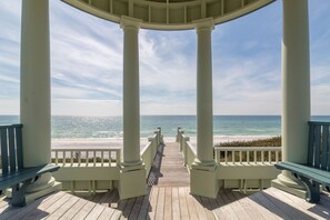 Una terraza cubierta de madera con columnas blancas y bancos, con vistas a una playa y al océano.
