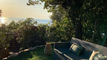 Un canapé en osier avec des coussins, une chaise en osier et une petite table ronde dans un jardin offrant une vue sur la mer.