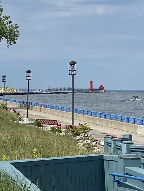 A coastal promenade with a red lighthouse, benches, and a blue railing. - GRAND LANDING CONDO UNIT 206, Outrageously Spacious, Sleeps 5-6, 3 BDR 2.5 BTH (Grand Haven)