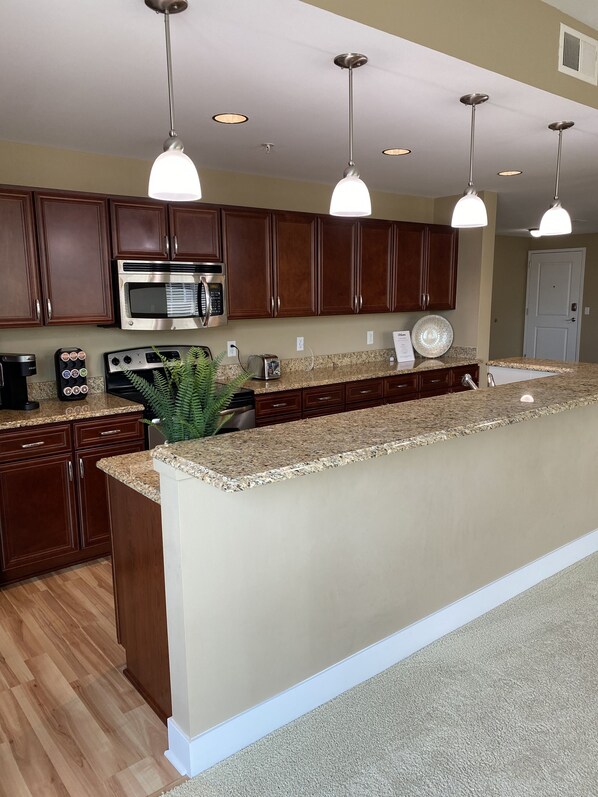 A modern kitchen with dark wooden cabinets, a stainless steel microwave, a built-in oven, and a granite countertop. There are pendant lights above the island and a large plant on the counter. - GRAND LANDING CONDO UNIT 206, Outrageously Spacious, Sleeps 5-6, 3 BDR 2.5 BTH (Grand Haven)