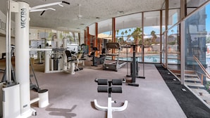 A modern gym with various exercise equipment, large windows, and a view of palm trees and a pool. - Posh Condo in Ocotillo Lodge in South PS (Palm Springs)
