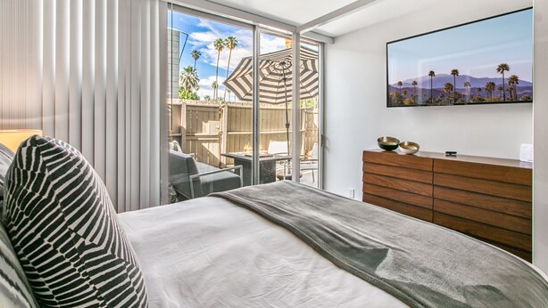 A hotel room with a bed, a TV, a wooden dresser, and a balcony with a view of palm trees and mountains. - Posh Condo in Ocotillo Lodge in South PS (Palm Springs)