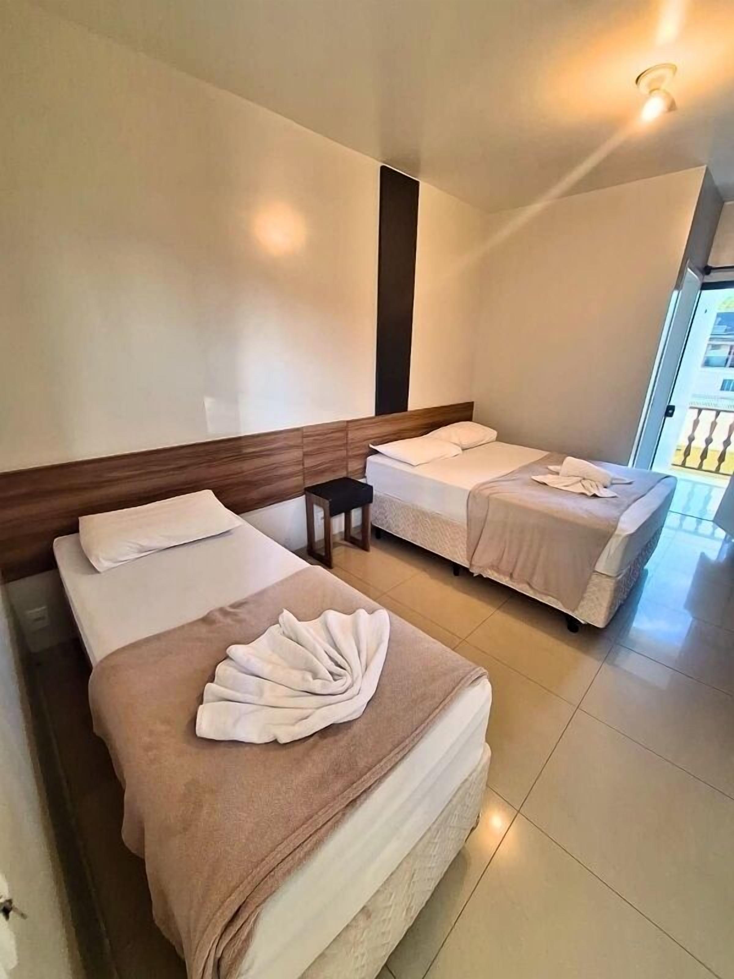 Quarto de hotel com duas camas, uma cabeceira de madeira, uma mesinha e vista para o exterior.