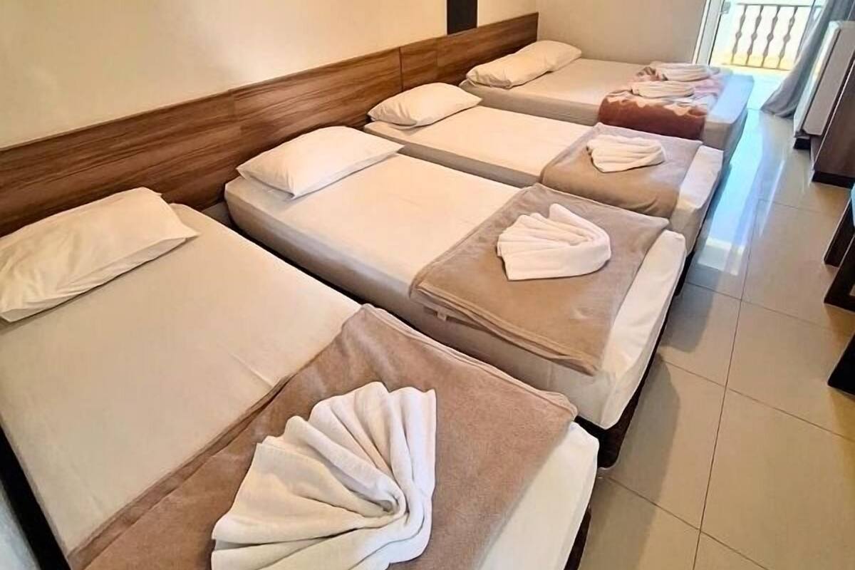 Quarto de hotel com quatro camas dispostas em linha, cada uma com cabeceira de madeira e roupa de cama branca.