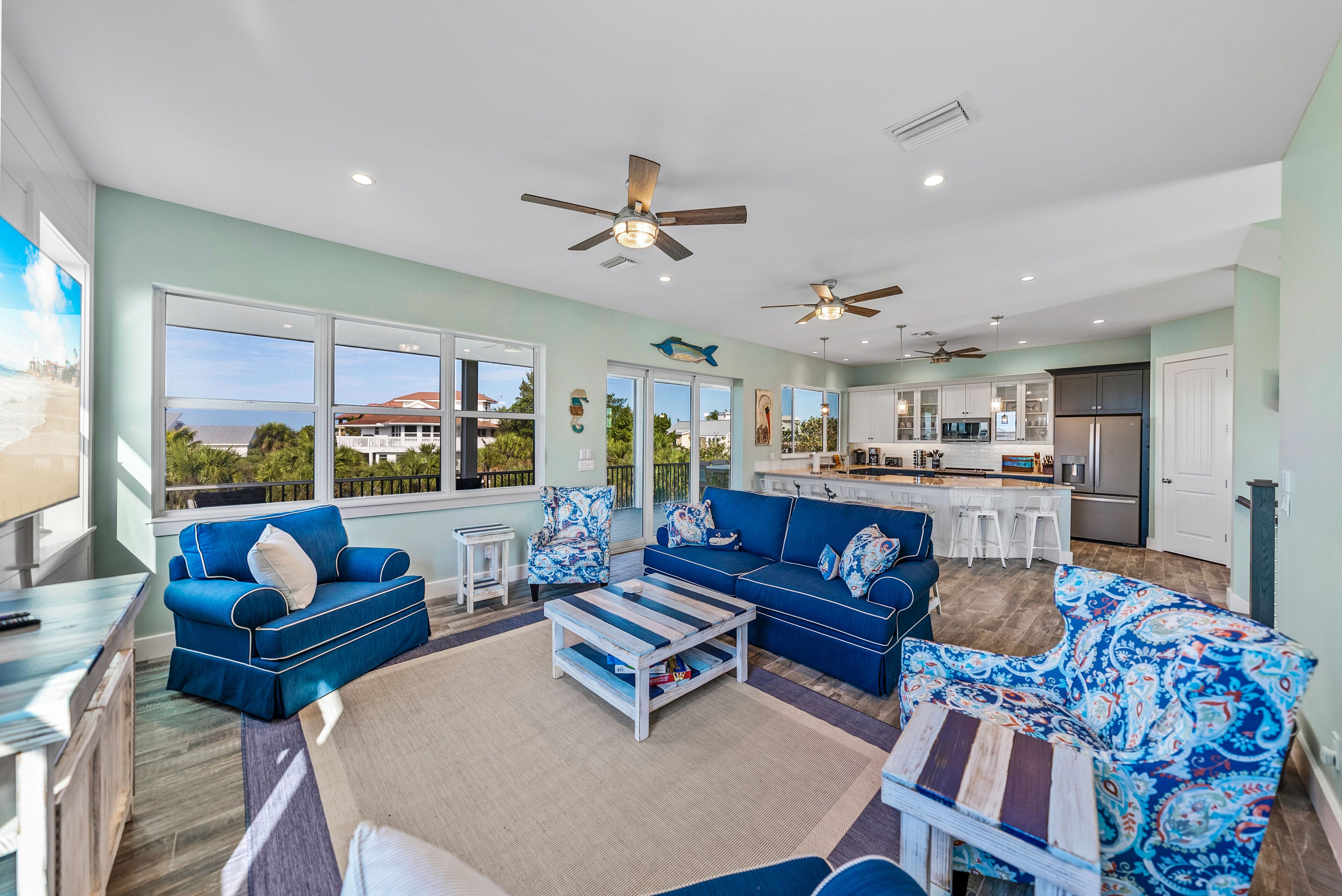 A spacious living room with blue sofas and a coffee table, large windows with a view, and a modern kitchen in the background.