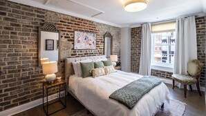 Een slaapkamer met een bakstenen muur, een groot bed met veel kussens, een nachtkastje met een lamp, een stoel en een raam met gordijnen.