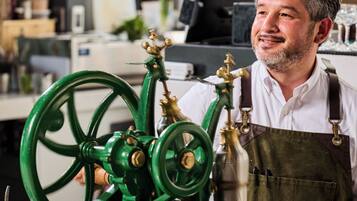 A person operating a green mechanical device, possibly a coffee grinder, in a kitchen setting with a 'SHASH' sign in the background.
