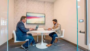 Two men seated at a round table in a modern office with patterned wallpaper and a large abstract painting on the wall.