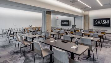 A conference room with multiple tables and chairs, a large screen displaying 'SHASHI HOTEL', and a carpeted floor.