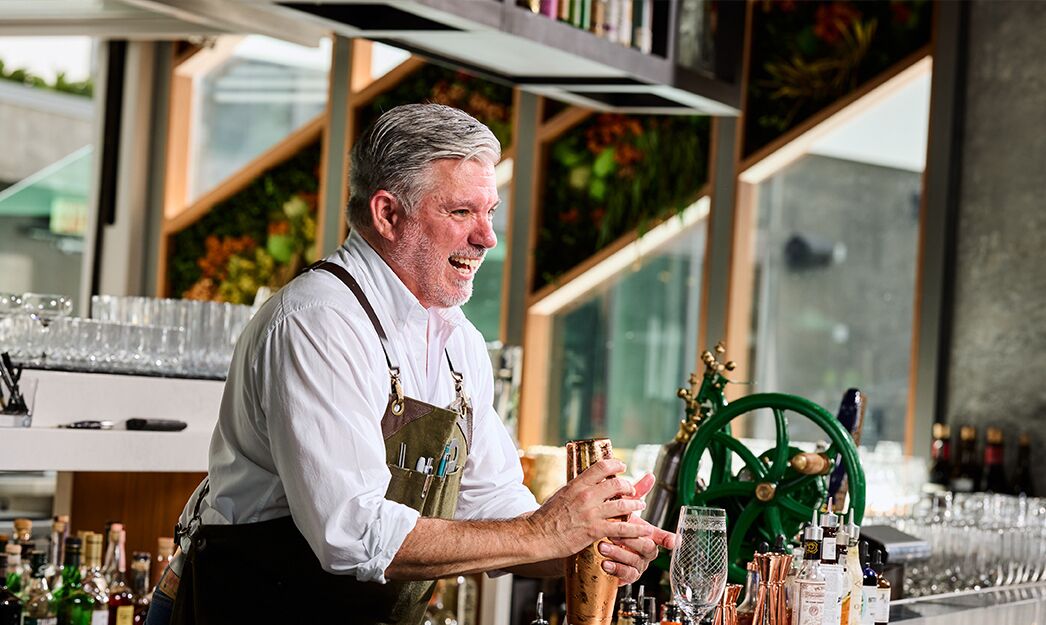 A bartender at a bar with a green wheel, various bottles, and glasses on the counter.