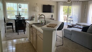 A modern kitchen with a central island, a dining area with a table and chairs, a grey sofa, and a flat-screen TV mounted on the wall. - Beautiful Second Floor 2-Bedroom Two-Bath Condo With Water and Golf Views (Calabash)