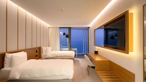 A modern hotel room with two beds, a desk, a TV, and a large window offering a view of the city. - Radisson Beach Resort Larnaca (Oroklini)