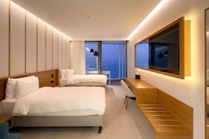 A modern hotel room with two beds, a desk, a TV, and a large window offering a view of the city.