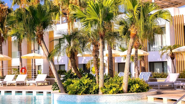 A swimming pool with lounge chairs and palm trees, surrounded by a modern building with large windows and a covered area. - Radisson Beach Resort Larnaca (Oroklini)