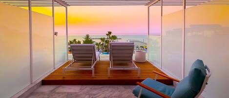 Un balcone con due poltrone lounge e un pavimento in legno, con vista sulla città e sul mare al tramonto.