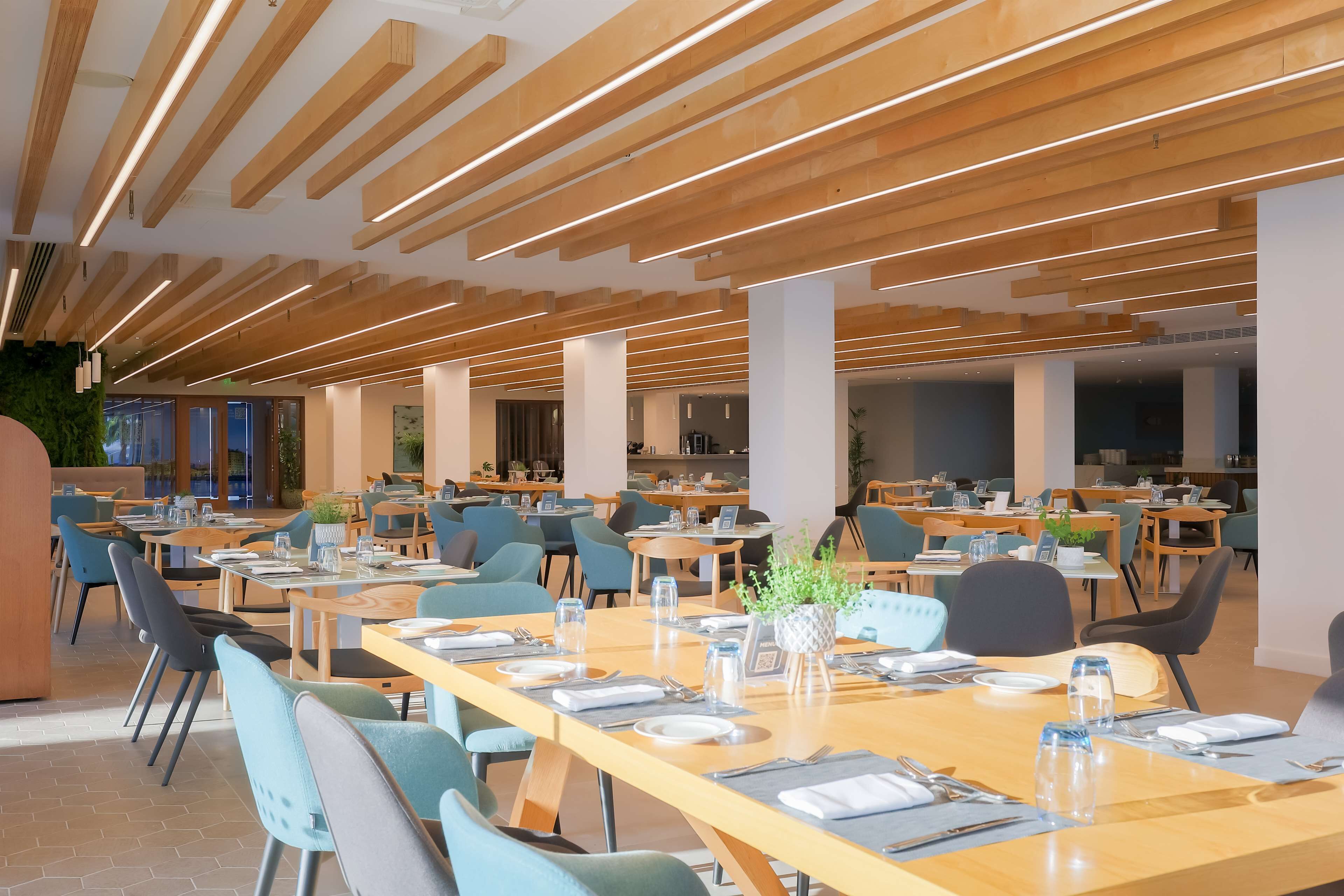 A spacious dining area with long wooden tables, blue chairs, and modern pendant lighting.