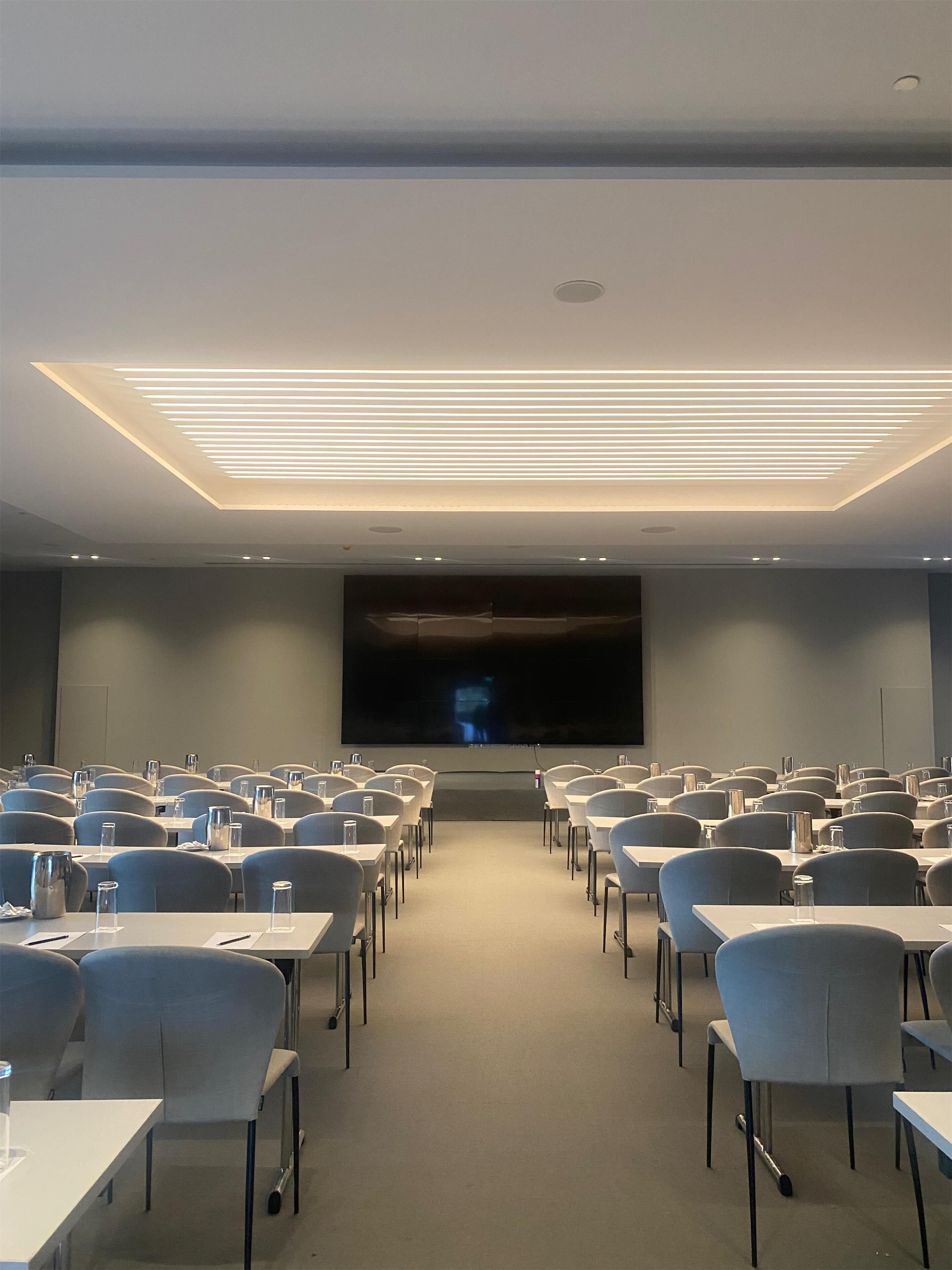 A conference room with rows of tables and chairs, a large screen at the front, and recessed ceiling lighting.
