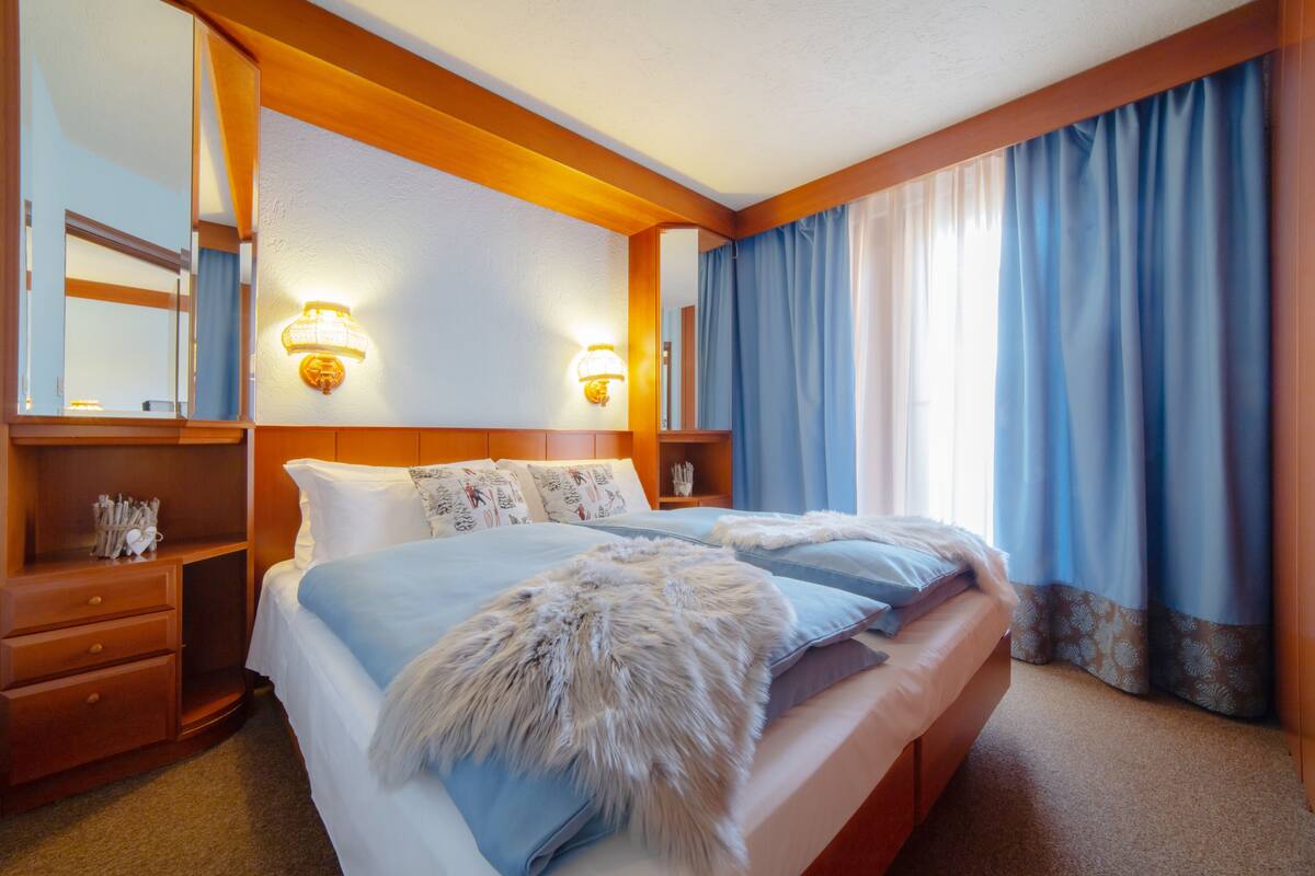 Ein Hotelzimmer mit einem großen Bett, einem Holzkopfstück, zwei Nachttischen, einem Schminktisch, einem Spiegel und einem Fenster mit blauen Vorhängen.