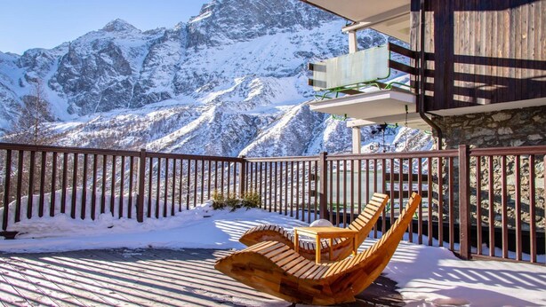 Eine Holzterrasse mit einem einzigartigen Stuhldesign, einer Steinmauer und Blick auf schneebedeckte Berge.