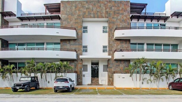 Un bâtiment moderne à deux étages, doté d’une entrée centrale, de balcons à chaque étage et d’un espace de stationnement avec des voitures.