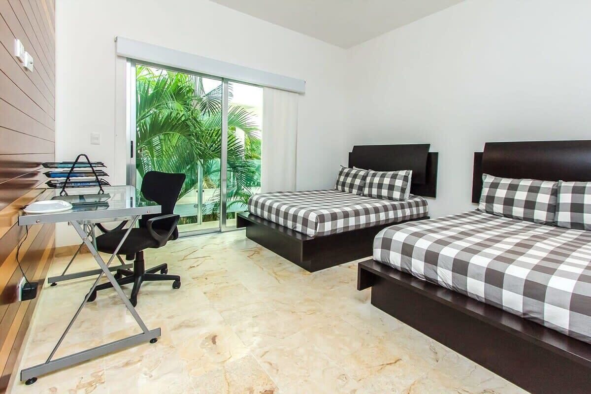 Quarto de hotel com escrivaninha, duas camas, uma cadeira e vista para o verde.