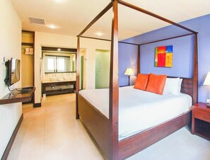 Una cama con dosel, ropa de cama blanca y almohadas naranjas, una mesita de noche con una lámpara y un baño con un espejo grande y lavamanos dobles.