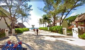 Un sentier menant à une plage, avec une structure à toit de chaume, entourée de verdure et sous un ciel dégagé.