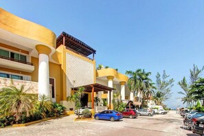 A hotel with a yellow facade, white columns, and a covered entrance. There are cars parked in front and palm trees along the street.