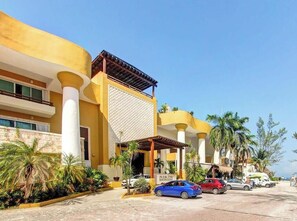 Un hotel con fachada amarilla, columnas blancas y un vestíbulo cubierto. Hay coches estacionados al frente y palmeras a lo largo de la calle.