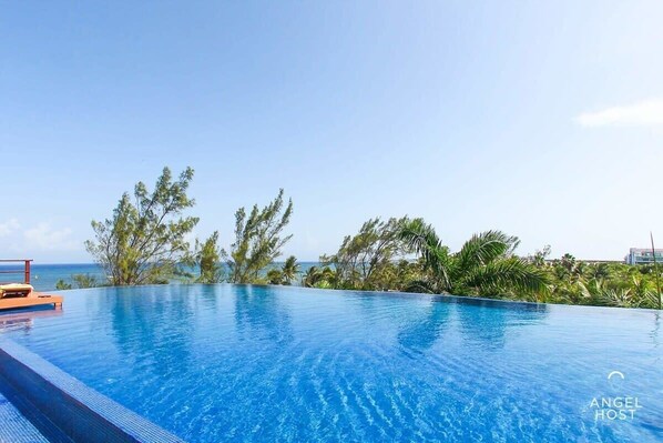 A clear blue infinity pool with a wooden deck, surrounded by tropical vegetation and a view of the ocean.