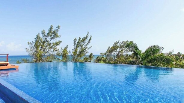 Une magnifique piscine à débordement d’un bleu profond, dotée d’une terrasse en bois, entourée d’une végétation tropicale et offrant une vue sur l’océan.