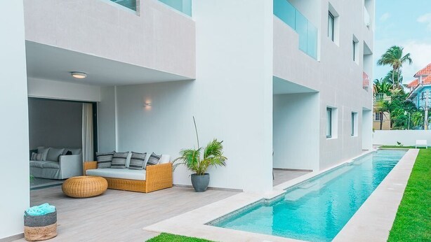Un edificio moderno con piscina, zona relax con cuscini e una pianta in vaso, e un prato ben curato.