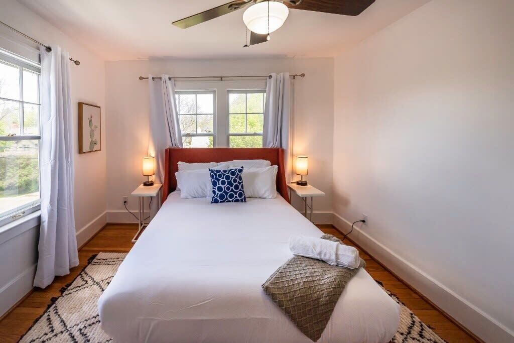 Une chambre à coucher avec un lit, deux lampes de chevet, une fenêtre avec des rideaux, un ventilateur de plafond et une petite table avec une lampe.