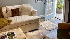 Ein Wohnzimmer mit einem beigen Sofa, einem Couchtisch mit Zeitschriften, einem gemusterten Teppich und einer Tür, die in einen Garten führt.