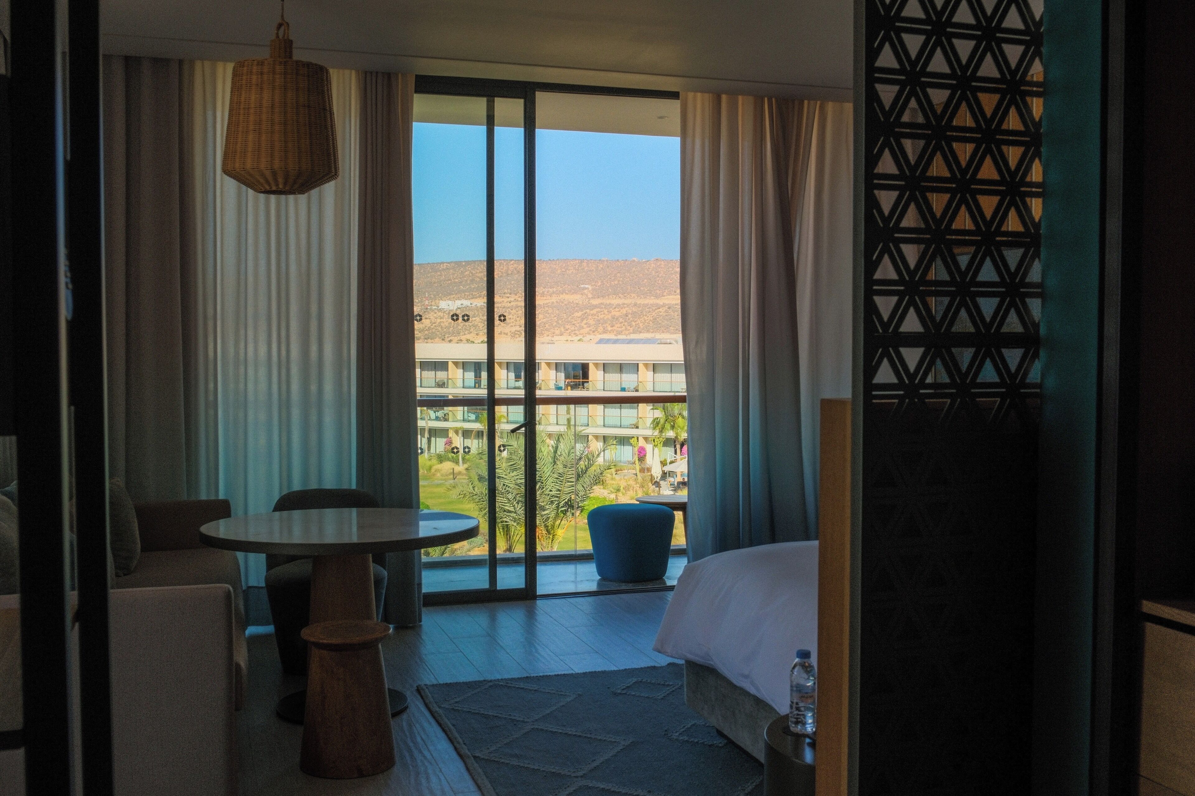 A hotel room with a large sliding glass door leading to a balcony, a round table, a sofa, and a bed.
