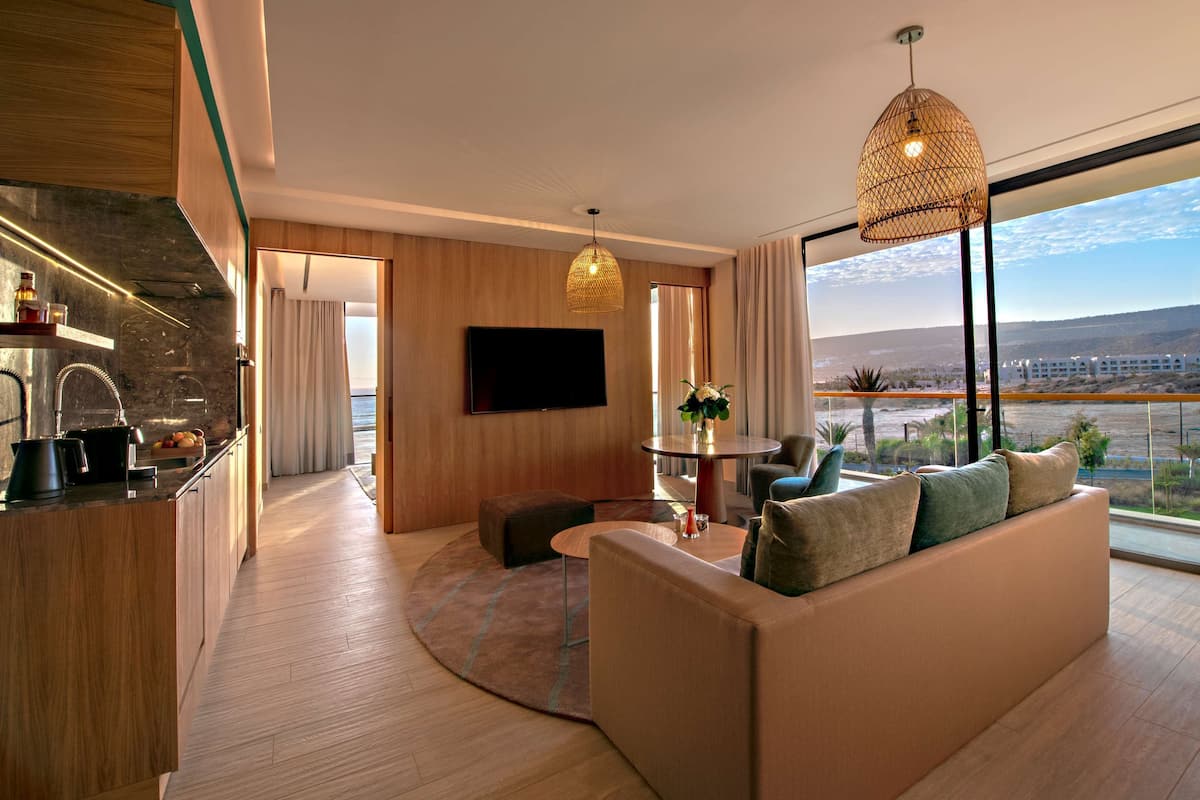 A modern living room with a sofa, a coffee table, and a TV. There is a kitchen area with a sink and a dining table. The room has large windows with a view of the city and hills.